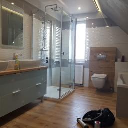 PRO-100 Krzysztof Gajewski - Wykończona łazienka na poddaszu z drewnopodobną podłogą, kabina prysznicowa ze szklanymi ściankami, umywalki nablatowe z szafką w kolorze błękitnym, wisząca toaleta i wanna.