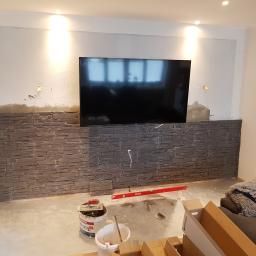 PRO-100 Krzysztof Gajewski - Montaż dekoracyjnej ściany z imitacji kamienia pod telewizorem, widoczne narzędzia, poziomica, wiadro z klejem i kartony z materiałem wykończeniowym.