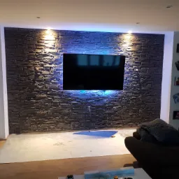 Ściana z dekoracyjnej cegły z podświetleniem LED za telewizorem w salonie, widoczne przygotowane podłoże pod podłogę.