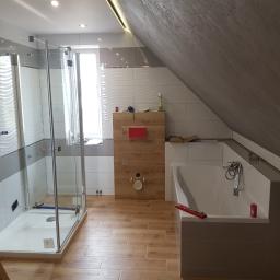 PRO-100 Krzysztof Gajewski - Wykończona łazienka na poddaszu z kabiną prysznicową, wanną, drewnopodobnymi płytkami na ścianie i podłodze oraz skośnym sufitem z oświetleniem LED.