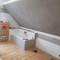 PRO-100 Krzysztof Gajewski - Ukończona łazienka na poddaszu z wanną obudowaną szarymi płytkami, drewnopodobną podłogą, szarą ścianą ze skosem i oświetleniem LED podsufitowym; widoczne narzędzia i akcesoria łazienkowe.