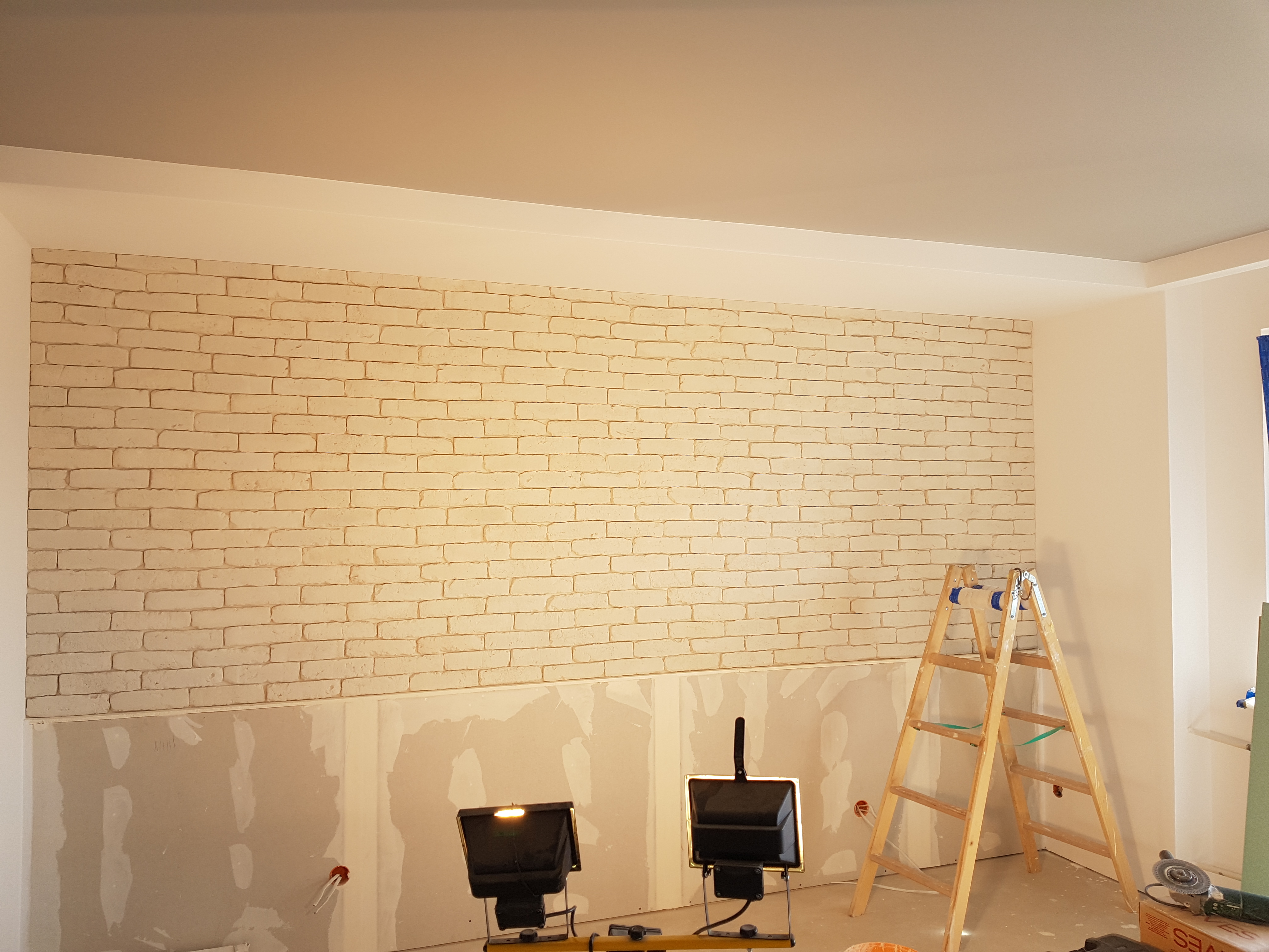 Ściana z imitacji cegły, częściowo wykończona, z widocznymi płytami gipsowo-kartonowymi poniżej, oświetlona lampami budowlanymi i z drewnianą drabiną w tle.