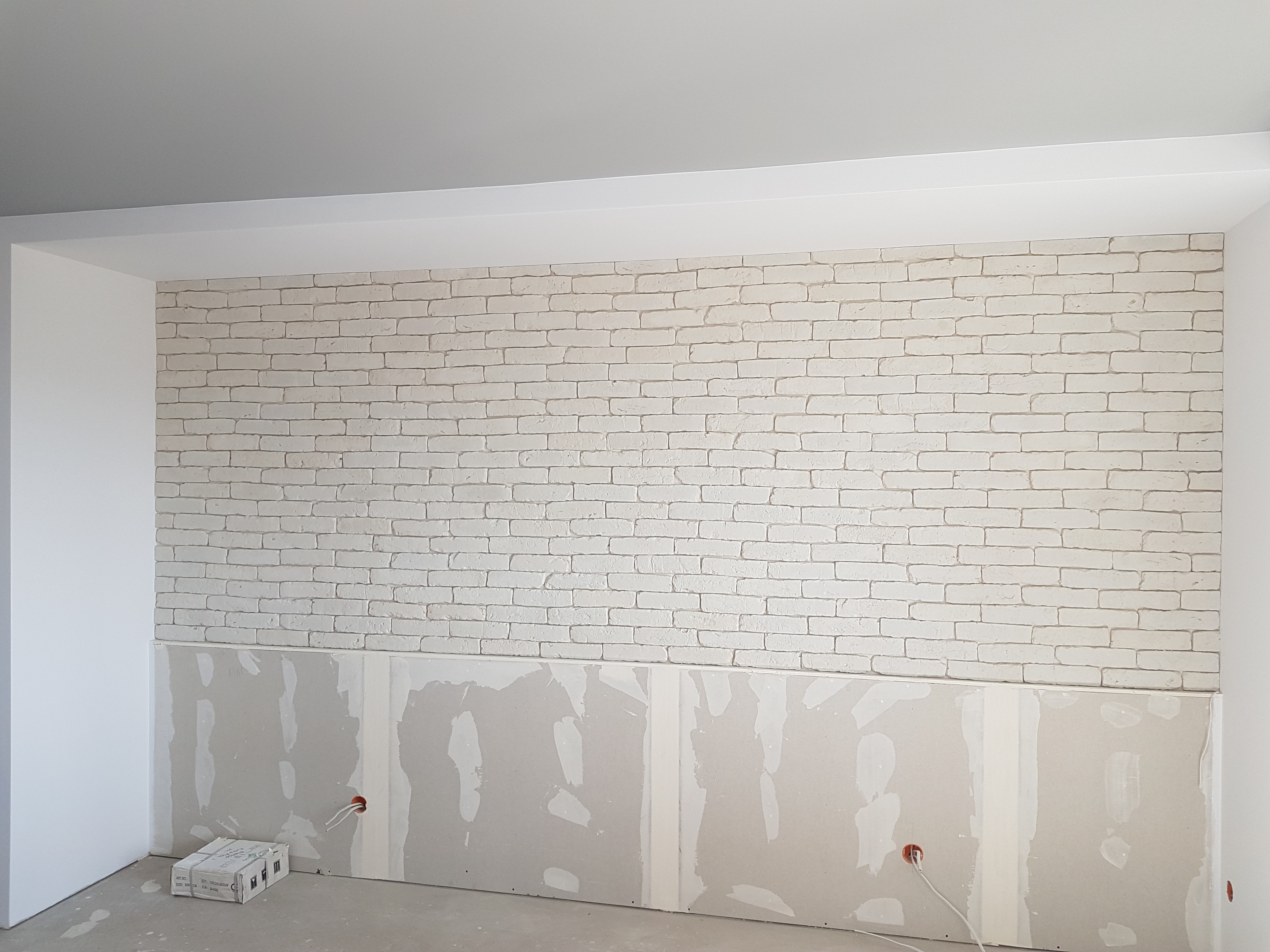 Ściana z cegieł pomalowanych na biało, z widocznymi fragmentami płyt na dole ściany i wystającymi kablami elektrycznymi.