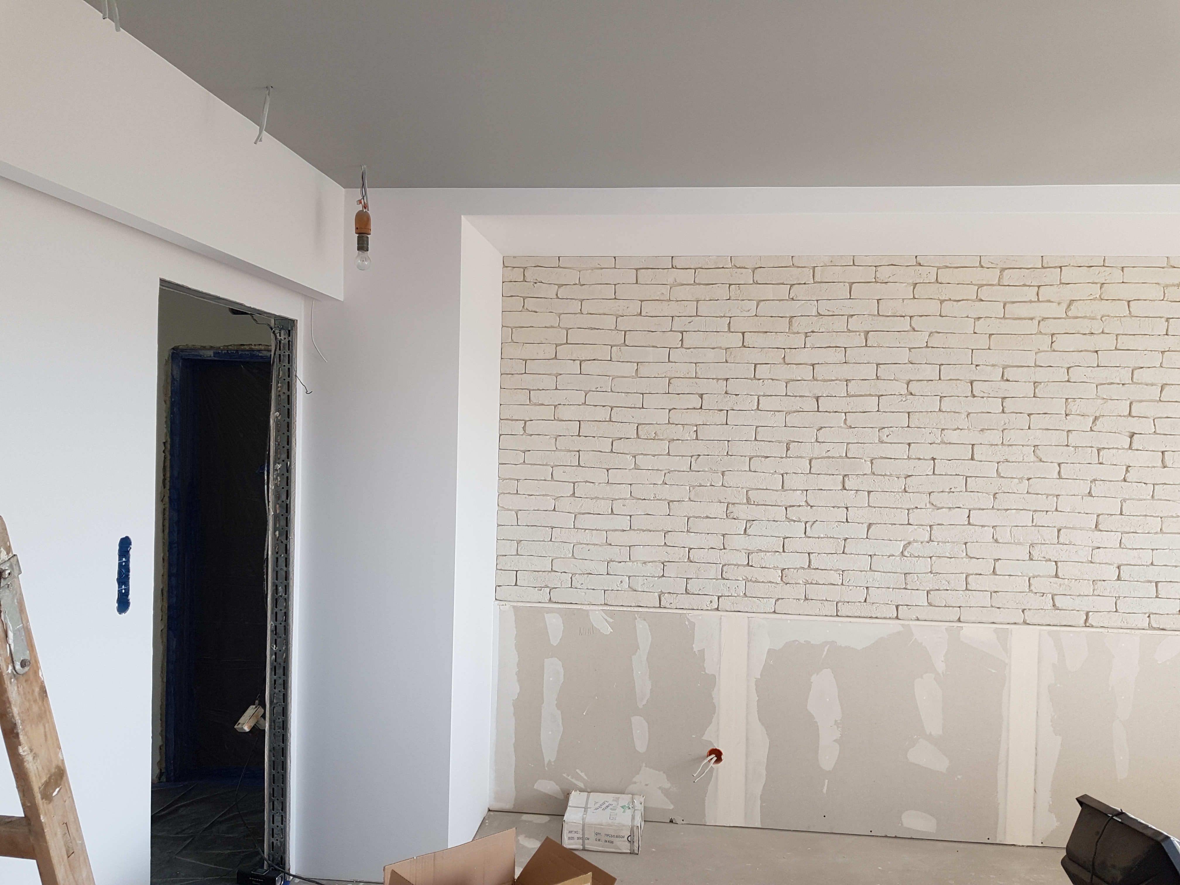 Wnętrze w trakcie wykańczania: białe ściany, imitacja cegły na górze, płyty na dole, widoczne przewody elektryczne, drabina i kartony na podłodze.