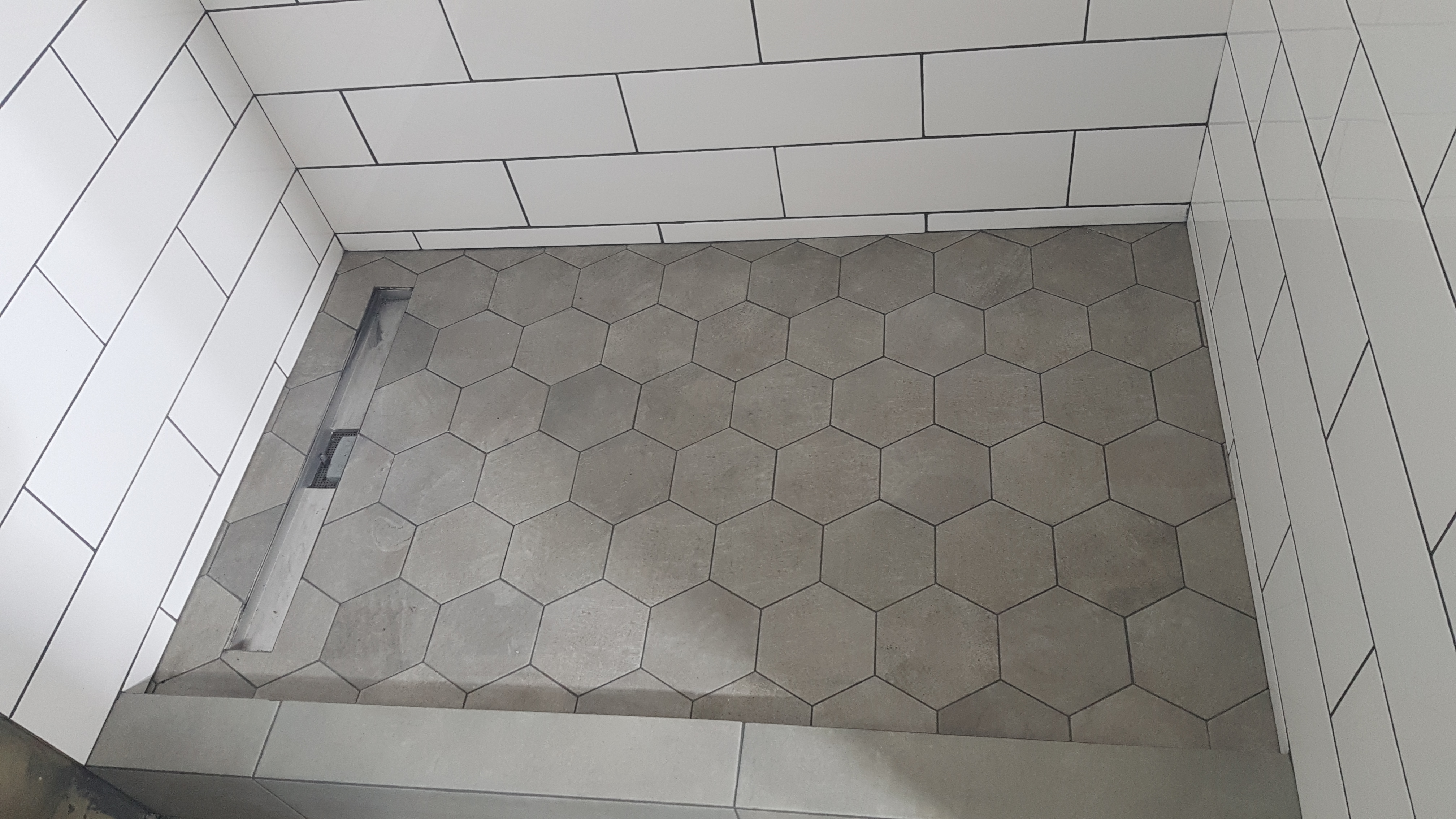 Wykończony brodzik z szarymi heksagonalnymi płytkami podłogowymi i białymi prostokątnymi płytkami ściennymi z ciemną fugą oraz odpływem liniowym