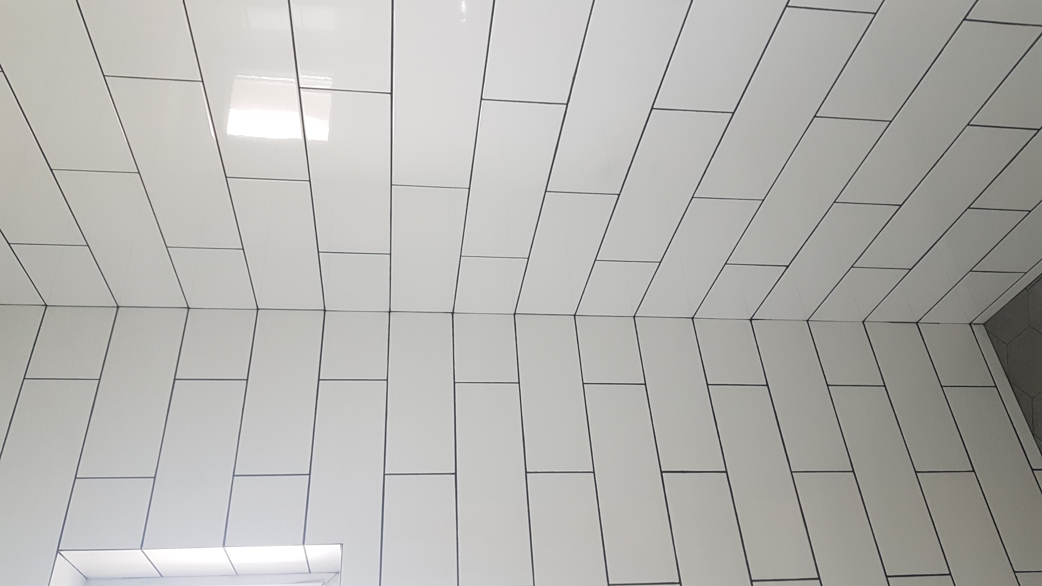 Białe, prostokątne płytki ułożone w cegiełkę na ścianie, z widoczną fugą w kolorze ciemnoszarym, z fragmentem heksagonalnych płytek w rogu.