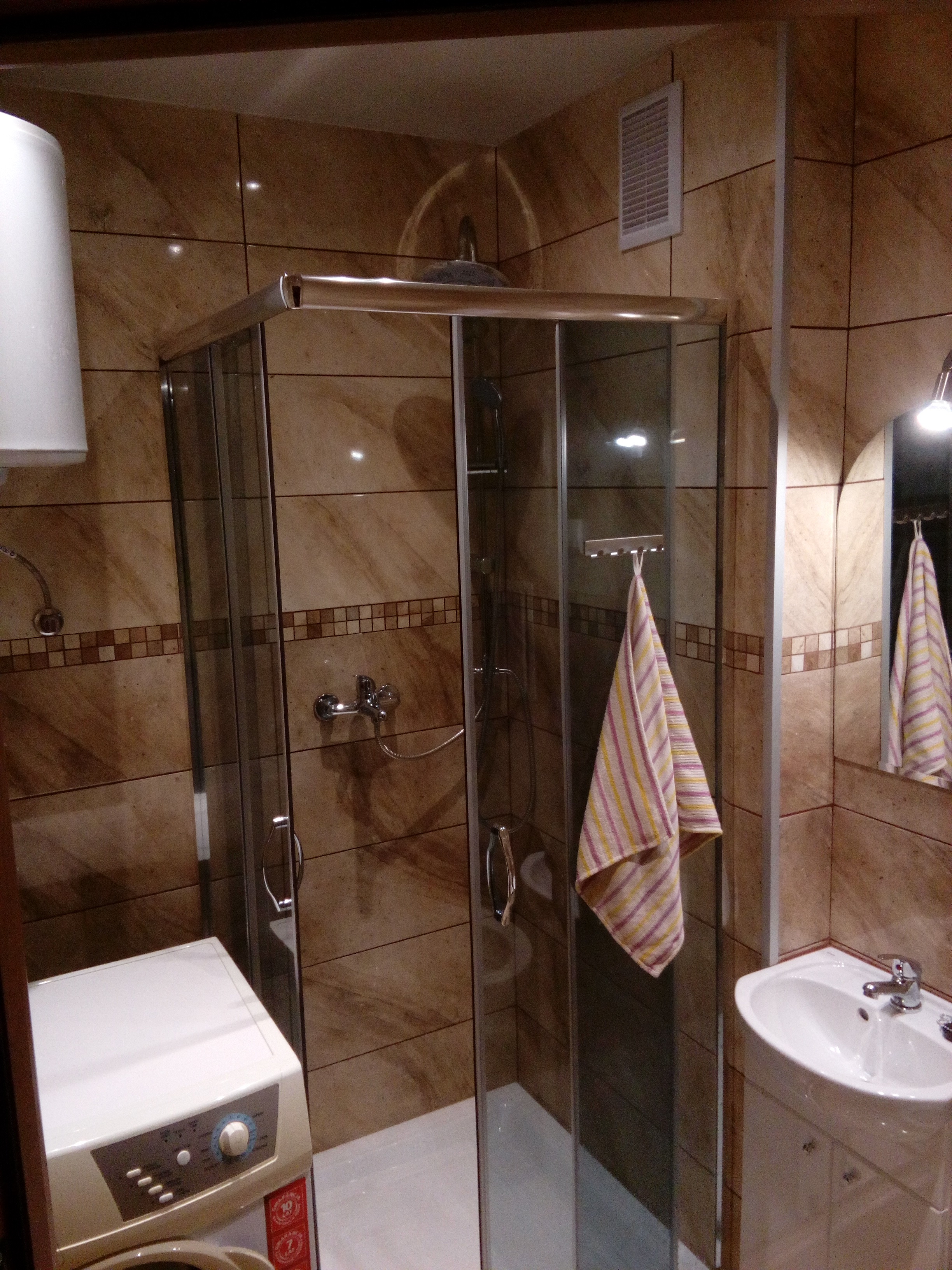 Ukończona łazienka z kabiną prysznicową, umywalką z szafką i pralką, wykończona brązowymi płytkami z dekoracyjnym paskiem, z ręcznikiem w paski zawieszonym na kabinie.