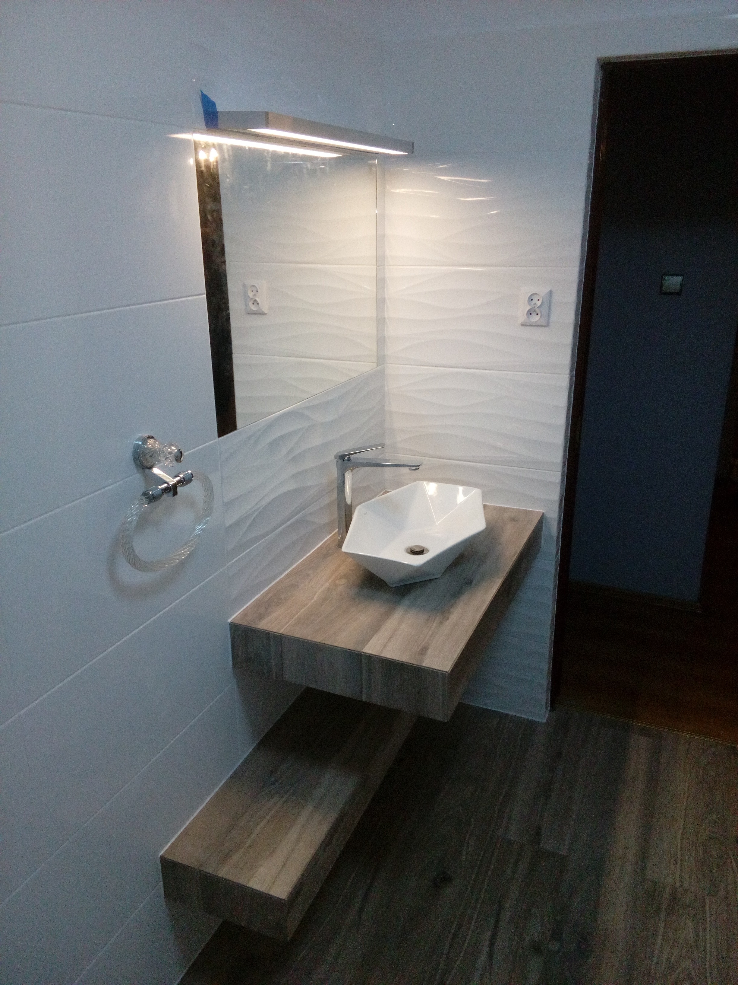 Nowoczesna łazienka z białą, geometryczną umywalką nablatową, drewnianym blatem i podświetlanym lustrem, uzupełniona o białe płytki z falistym wzorem i designerski uchwyt na ręczniki.