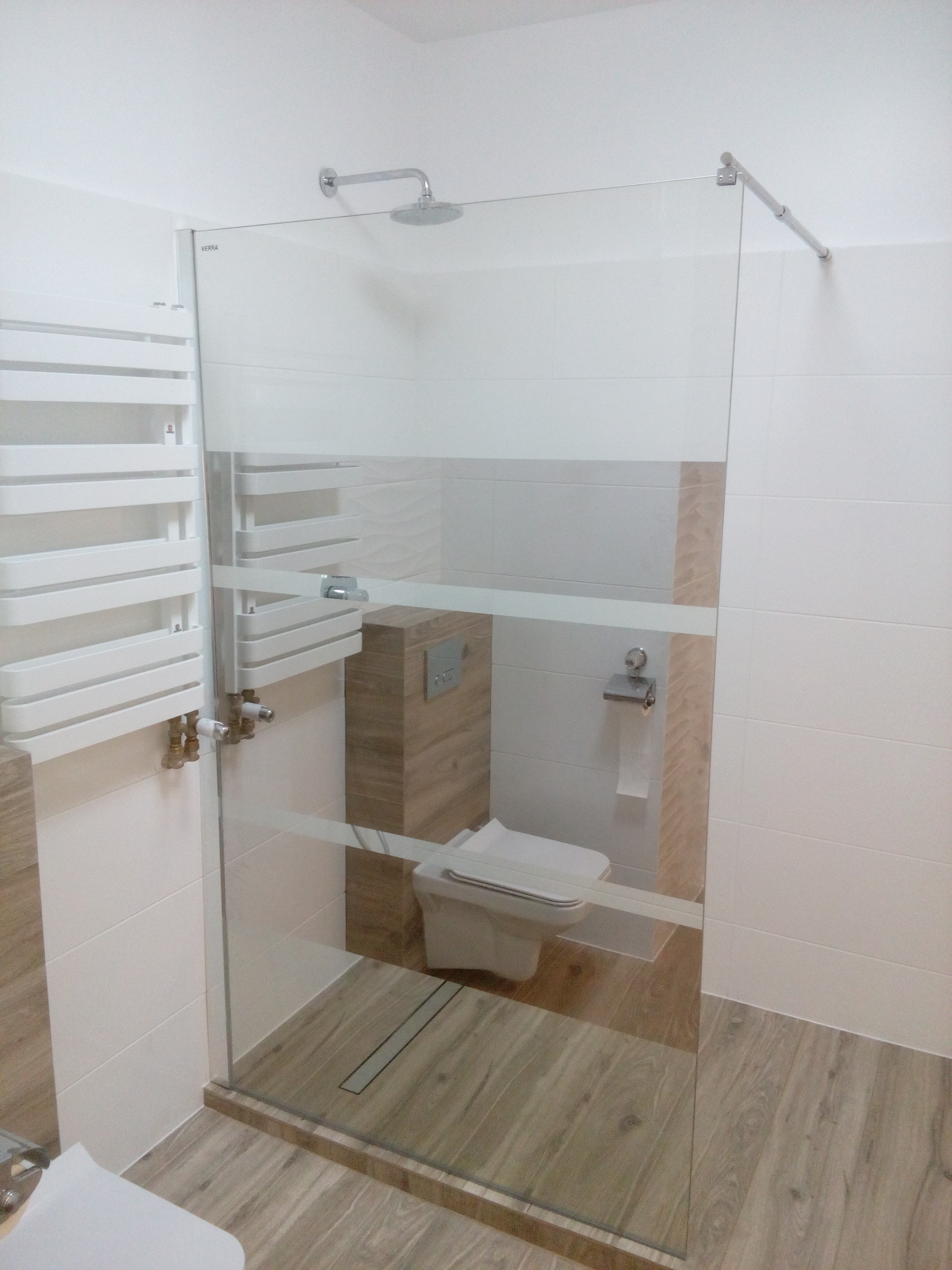 Nowoczesna łazienka po remoncie z kabiną prysznicową ze szklaną ścianką z poziomym pasem matowego szkła, grzejnikiem drabinkowym, toaletą podwieszaną i podłogą imitującą drewno.