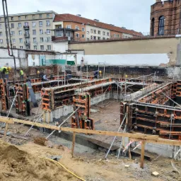 Fundamenty w trakcie budowy we Wrocławiu, z widocznymi deskowaniami i pracownikami w żółtych kaskach, na tle zabudowy miejskiej.