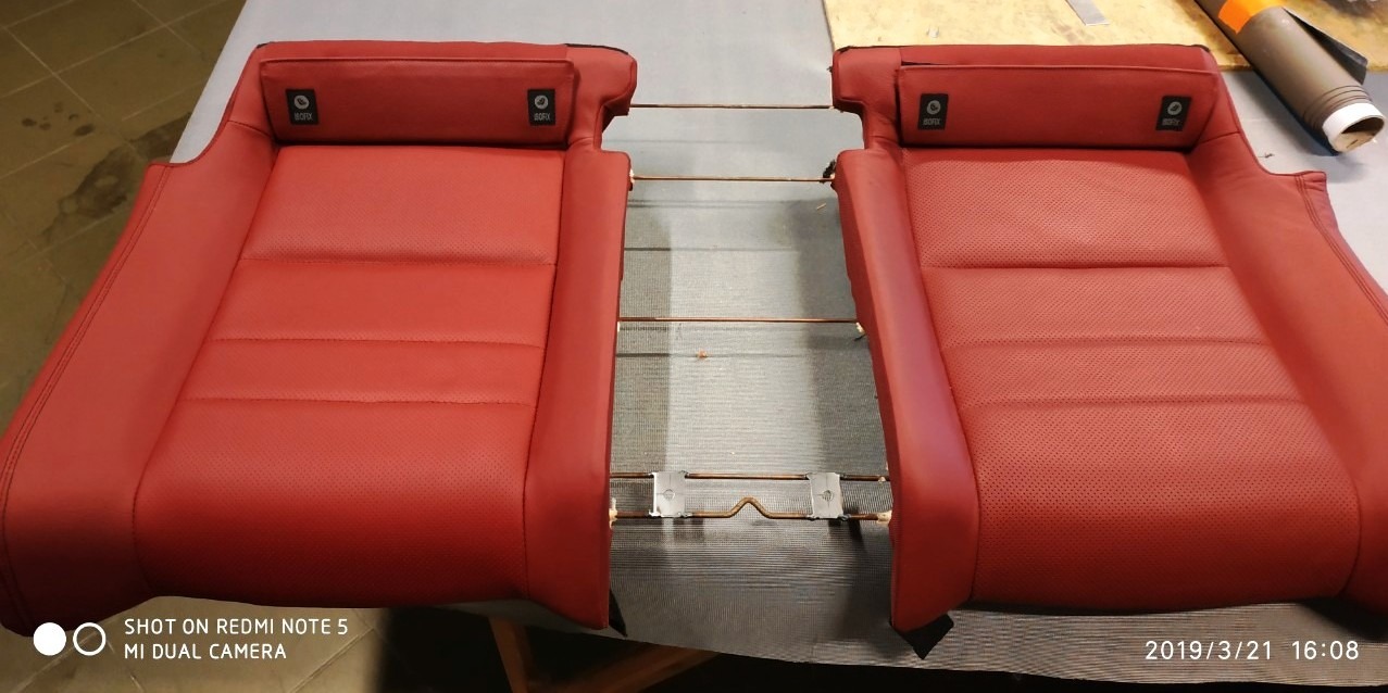 Dwa czerwone, skórzane fotele samochodowe z widocznymi metalowymi elementami konstrukcyjnymi i mocowaniami ISOFIX, leżące na warsztatowym stole.