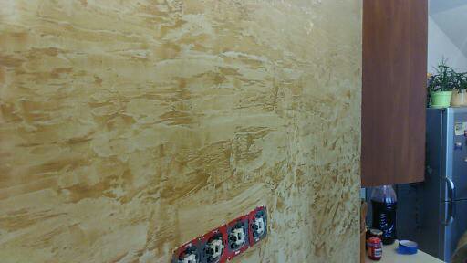 Ściana z dekoracyjną, beżowo-złotą fakturą, gniazdka elektryczne bez osłon, fragment szafki kuchennej i lodówki w tle.