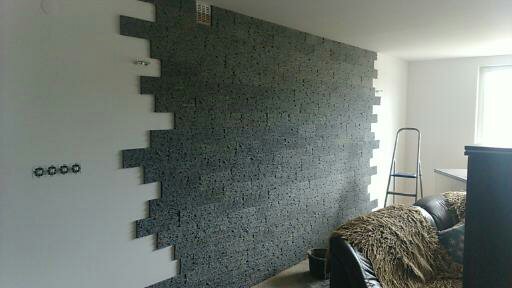 Częściowo wykończona ściana z dekoracyjnej, szarej cegły w pokoju, widoczna instalacja elektryczna, drabina oparta o ścianę, sofa w tle.