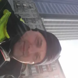 Autoportret mężczyzny w czapce i jaskrawej kamizelce odblaskowej na tle wysokiego budynku biurowego w słoneczny dzień.