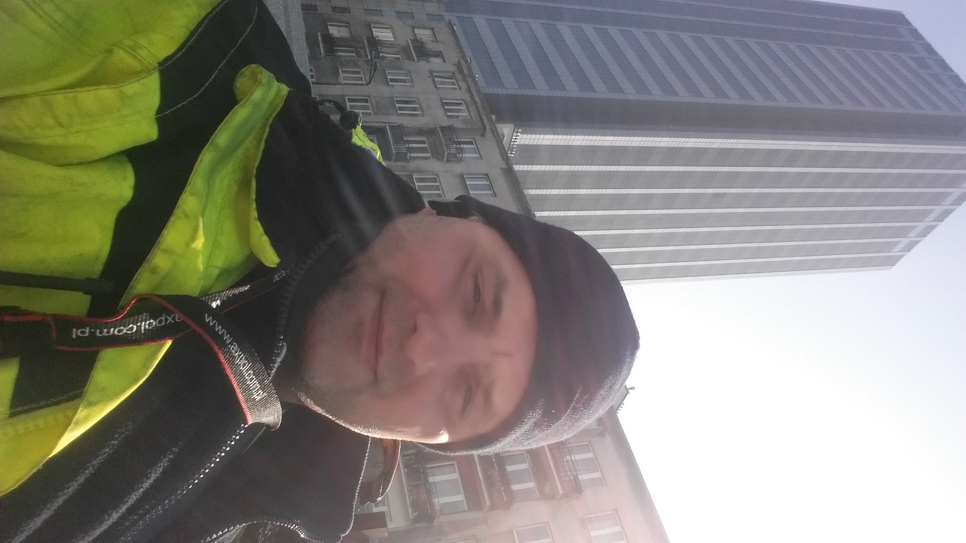 Autoportret mężczyzny w czapce i jaskrawej kamizelce odblaskowej na tle wysokiego budynku biurowego w słoneczny dzień.