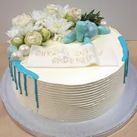 Tort urodzinowy z białym kremem, dekorowany białymi kwiatami, turkusową figurką żółwia i spływającym turkusowym lukrem, z napisem na białej tabliczce.