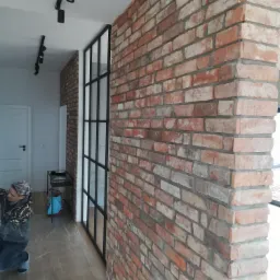 Ceglana ściana w odcieniach czerwieni i bieli, oddzielona czarną, metalową ramą z szybami od jasnego pomieszczenia z oświetleniem szynowym. Na podłodze folia ochronna, a obok pracownik w trakcie...