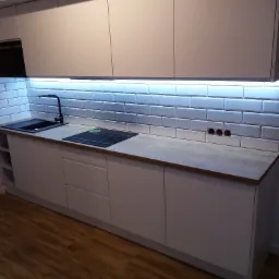 Nowoczesna kuchnia z białymi szafkami, drewnianym blatem, czarnym zlewem i płytą indukcyjną, podświetlana listwą LED pod szafkami wiszącymi, oraz białymi płytkami typu metro na ścianie.