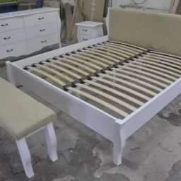 Białe łóżko z beżowym, pikowanym zagłówkiem i drewnianym stelażem, obok biała komoda, szafka nocna i tapicerowany puf z pikowaniem w romby.