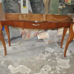 Lakierowany stolik z szufladami na giętych nogach w warsztacie stolarskim, widoczne niedoskonałości podłoża i materiały stolarskie w tle.