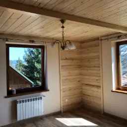 Pokój z drewnianą boazerią na ścianach i suficie, widok na góry przez okno, drewniana podłoga, żyrandol w stylu retro.