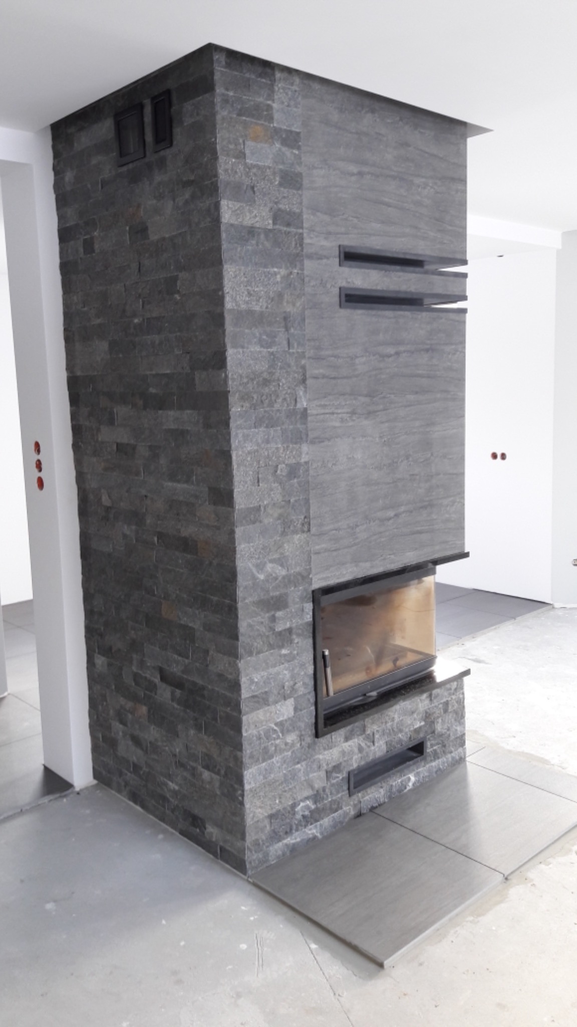 Nowoczesny kominek obłożony szarą, łupkową okładziną w surowym wnętrzu, z widocznymi elementami instalacji elektrycznej na ścianie.