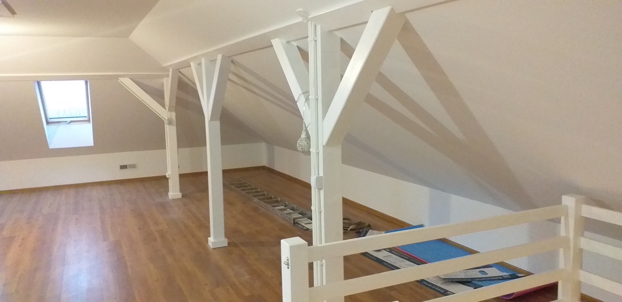 Poddasze z drewnianą podłogą i białymi, drewnianymi słupami konstrukcyjnymi. Widoczne okno dachowe i drabina leżąca na podłodze. Częściowo widoczna biała balustrada.