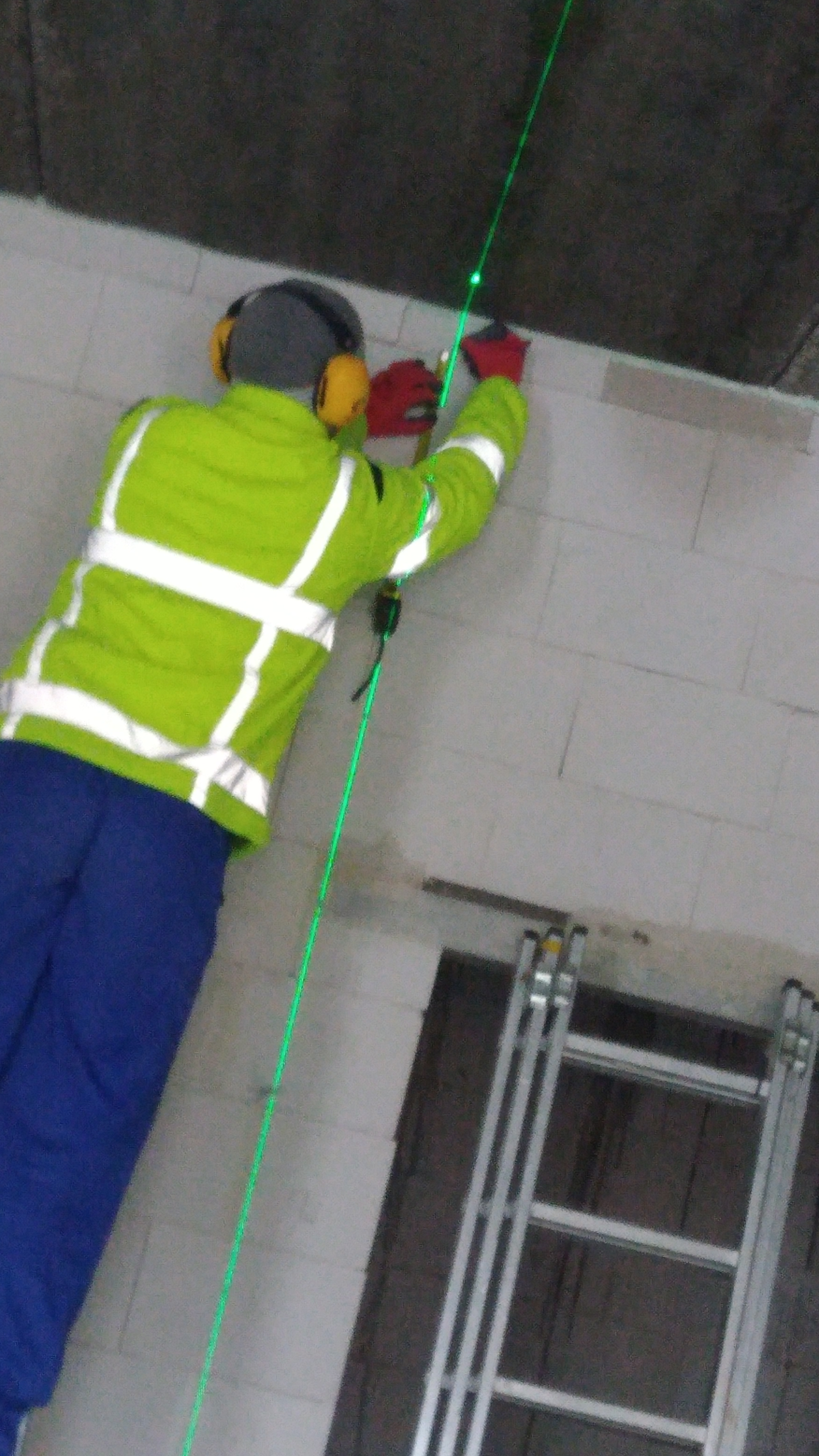 Pracownik w odblaskowej kurtce i ochronnikach słuchu, używa poziomicy laserowej do wyznaczania linii na ścianie z pustaków, widoczna drabina.