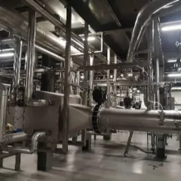 Rozbudowany system rur ze stali nierdzewnej w hali przemysłowej, liczne kolanka i łączenia, widoczne elementy mocujące, oświetlenie sufitowe.