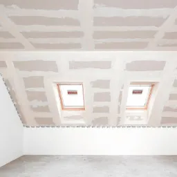 Poddasze z oknami dachowymi Velux w trakcie montażu płyt karton-gips, widoczne łączenia i szpachlowanie, zabezpieczone narożniki taśmą ochronną.