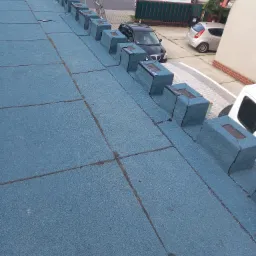 Niebieski dach pokryty papą z kominkami wentylacyjnymi, widok z góry na tle zaparkowanych samochodów i domów.