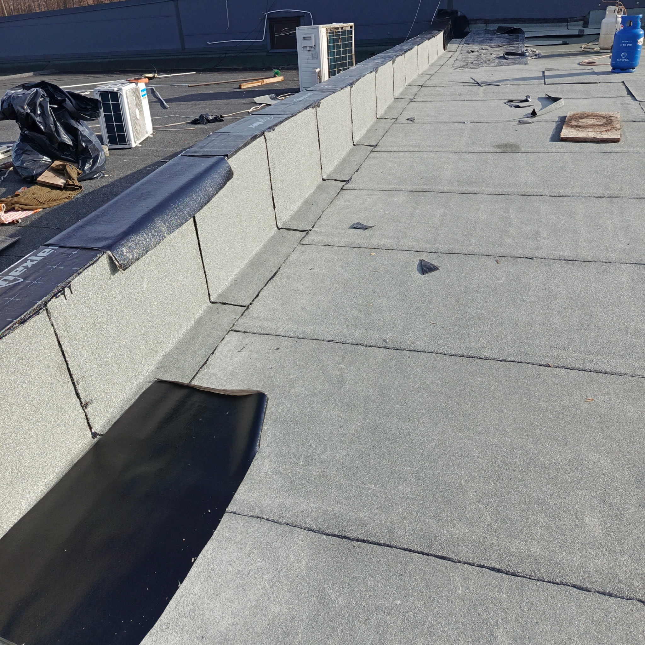 Płaski dach pokryty papą termozgrzewalną w trakcie prac, widoczne fragmenty papy, butle gazowe, klimatyzatory i narzędzia.