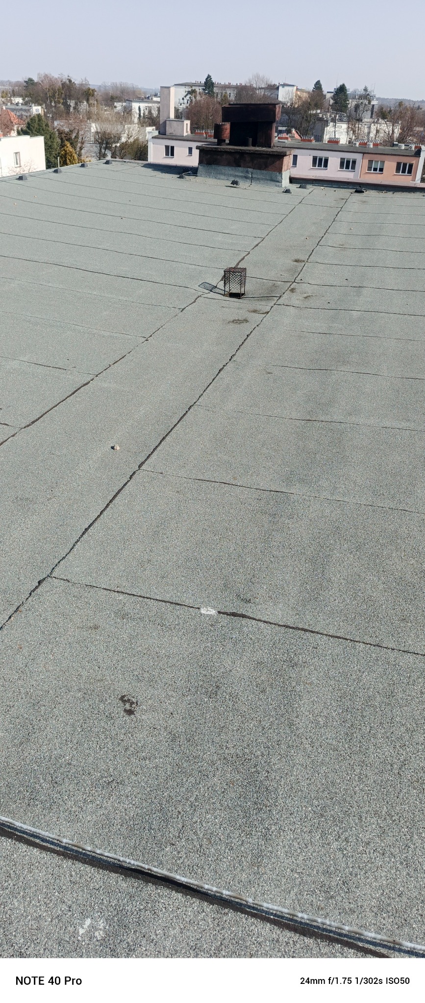 Szary dach pokryty papą termozgrzewalną z widoczną kratką wentylacyjną i budynkami w tle. Widok z góry, oświetlenie dzienne.