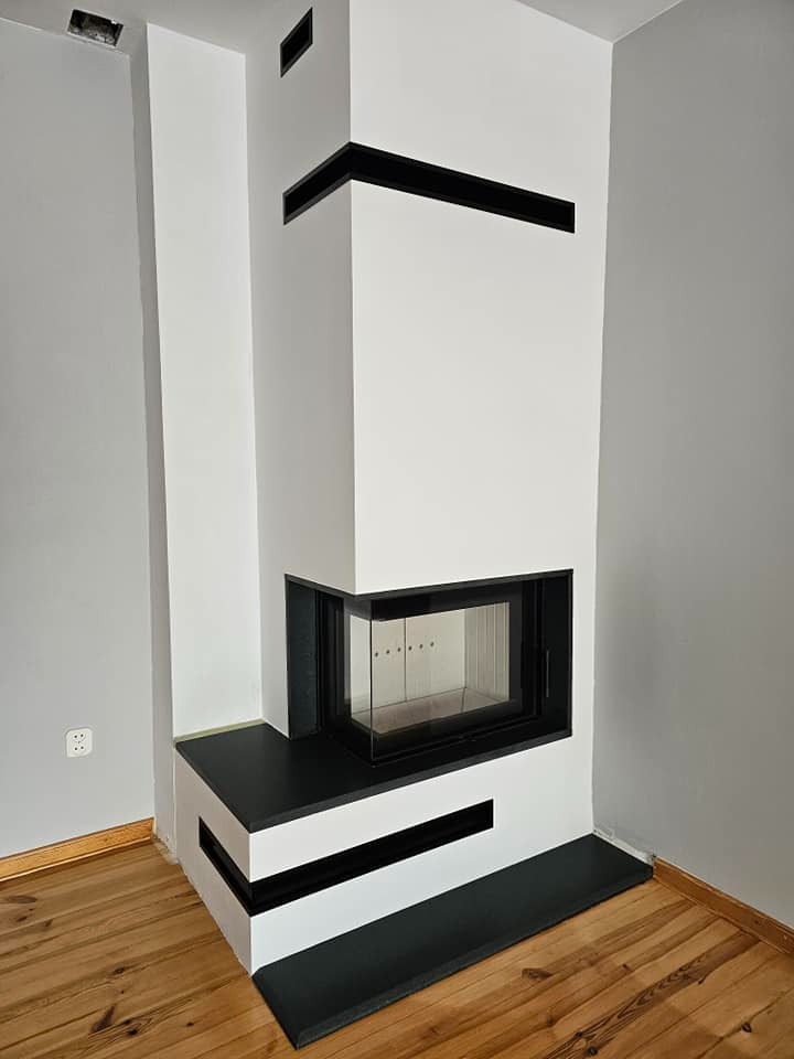 Nowoczesny, narożny wkład kominkowy z czarnymi akcentami, w minimalistycznym wnętrzu z drewnianą podłogą i szarą ścianą. Widok z boku na gotowy kominek.