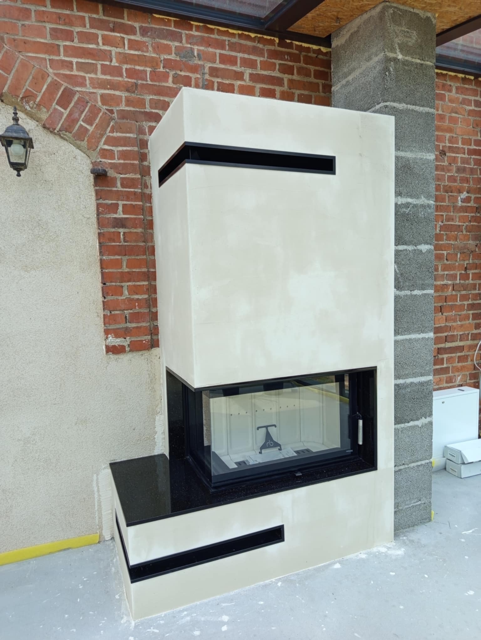 Nowoczesny, obudowany kominek w jasnej kolorystyce z czarnymi akcentami, widoczny na tle ceglanej ściany i betonowego filara. Minimalistyczny design.