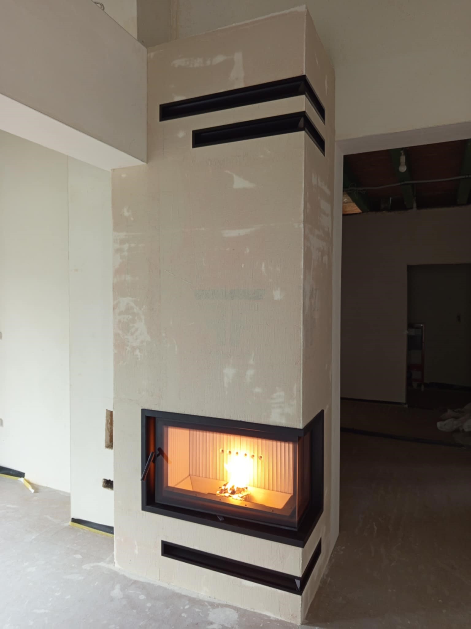 Nowoczesny kominek narożny z czarnymi akcentami, w surowym wnętrzu. Widoczne płomienie i niedokończone ściany.