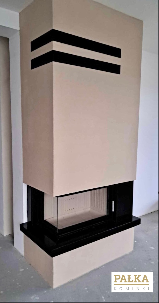 Nowoczesny kominek z beżową obudową i czarnymi akcentami, widoczne wnętrze paleniska z jasną wyściółką. Minimalistyczny design w surowym wnętrzu.