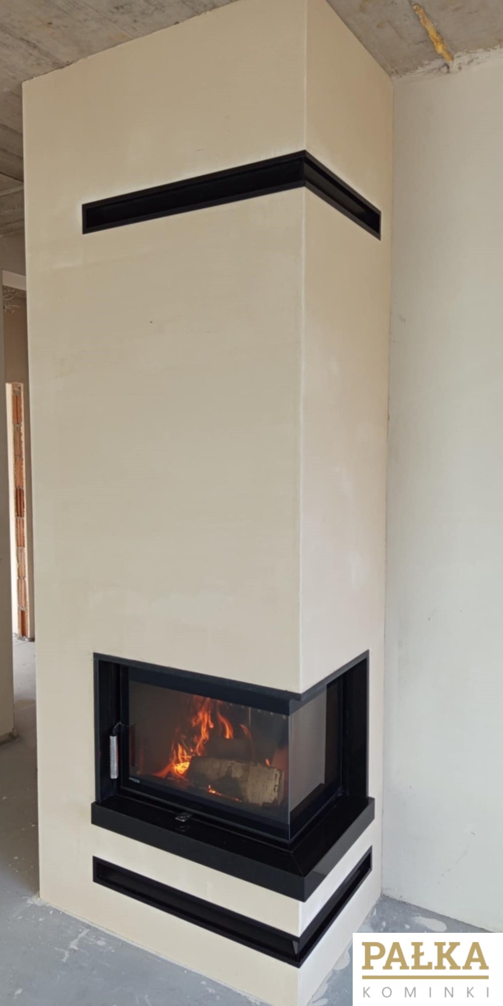 Narożny kominek z czarnymi elementami wykończeniowymi, widoczny płomień, surowy betonowy strop w tle, logo firmy w prawym dolnym rogu.