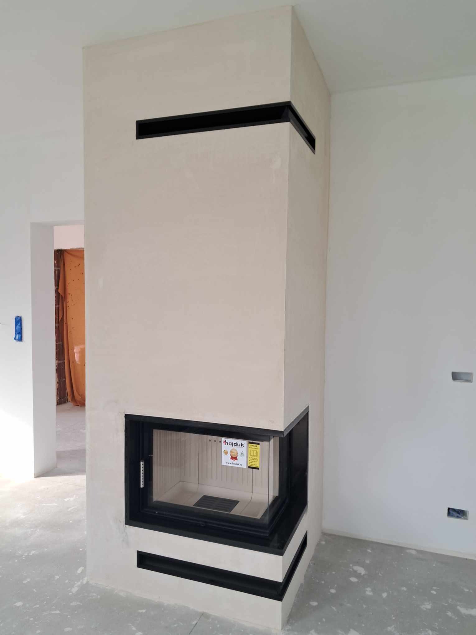 Nowoczesny kominek Hajduk z czarnym obramowaniem i jasnym wykończeniem, w surowym wnętrzu, przed oddaniem do użytku, z widoczną naklejką informacyjną na szybie.