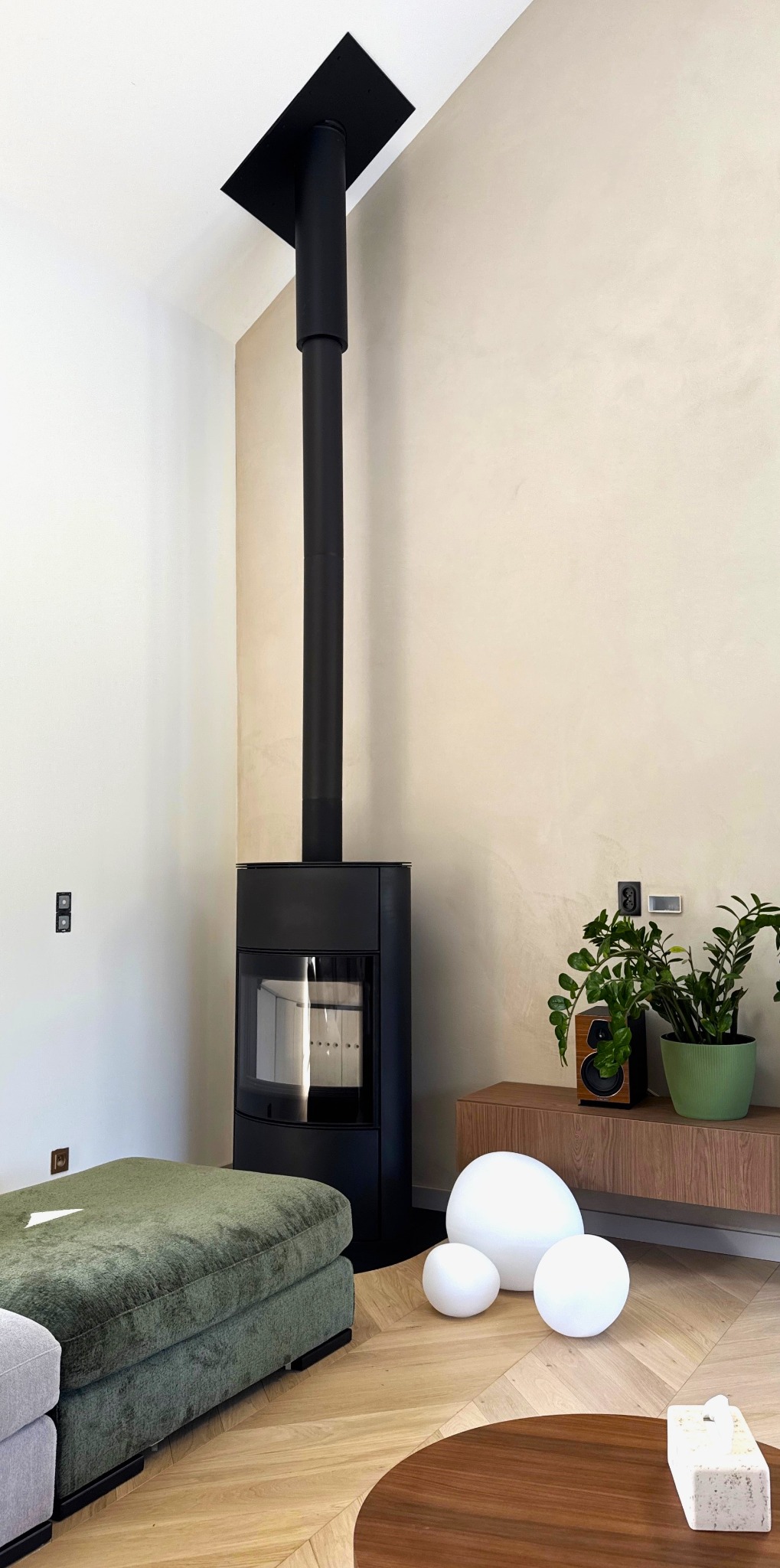 Nowoczesny, czarny kominek wolnostojący z długim przewodem kominowym w jasnym salonie. Obok pufa w odcieniach zieleni i dekoracyjne lampy kuliste na parkiecie.
