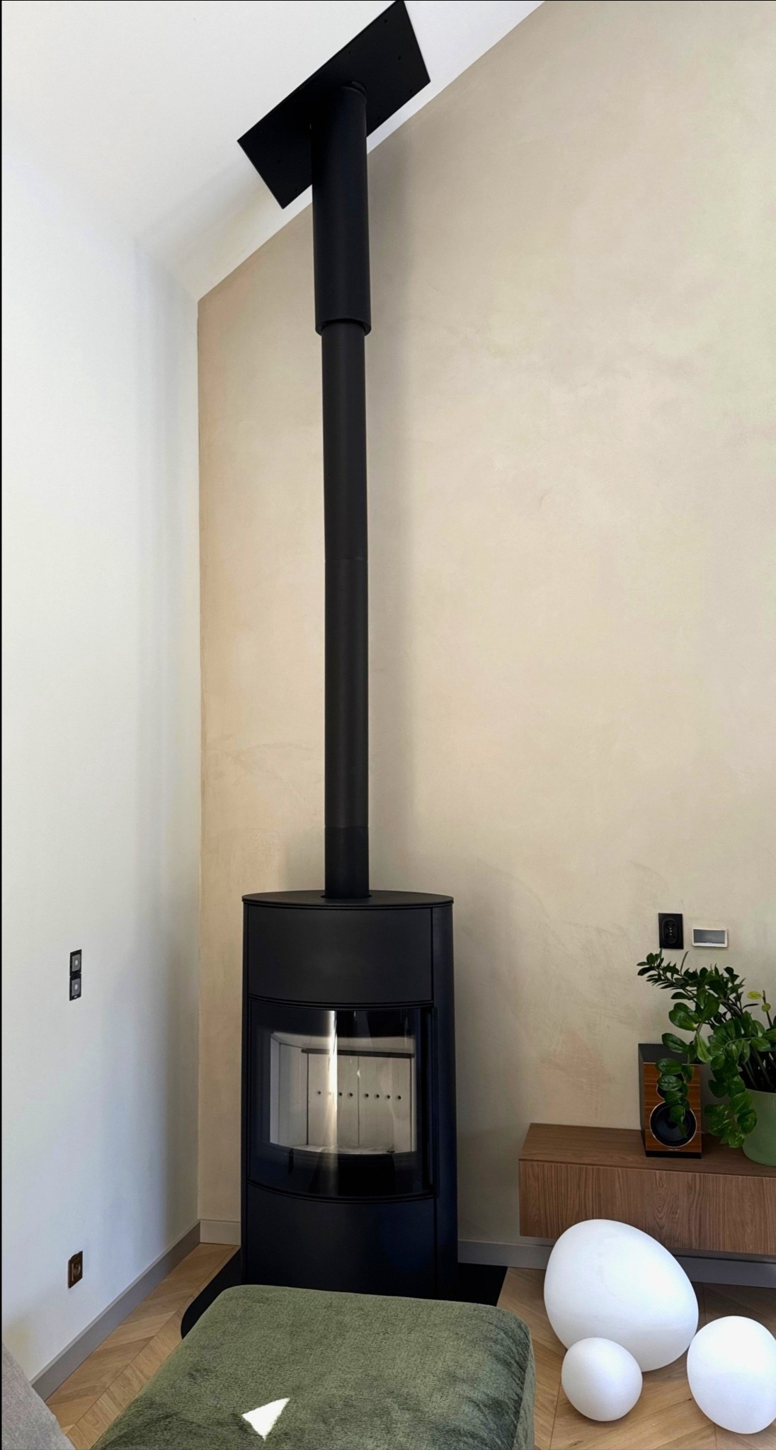 Nowoczesny, czarny kominek wolnostojący z długą rurą odprowadzającą dym, ustawiony w jasnym wnętrzu z drewnianą podłogą i elementami dekoracyjnymi.