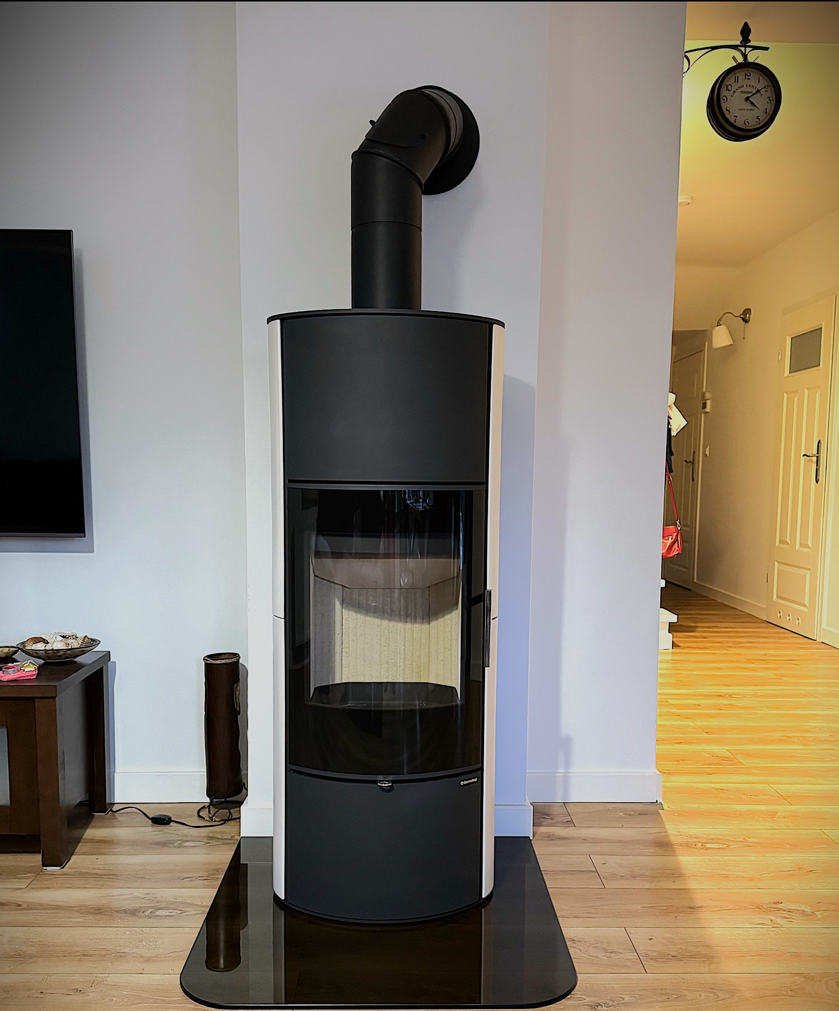 Nowoczesny, czarno-biały kominek wolnostojący z szybą, na czarnej szklanej płycie podłogowej. W tle jasne ściany, drewniana podłoga i zegar.