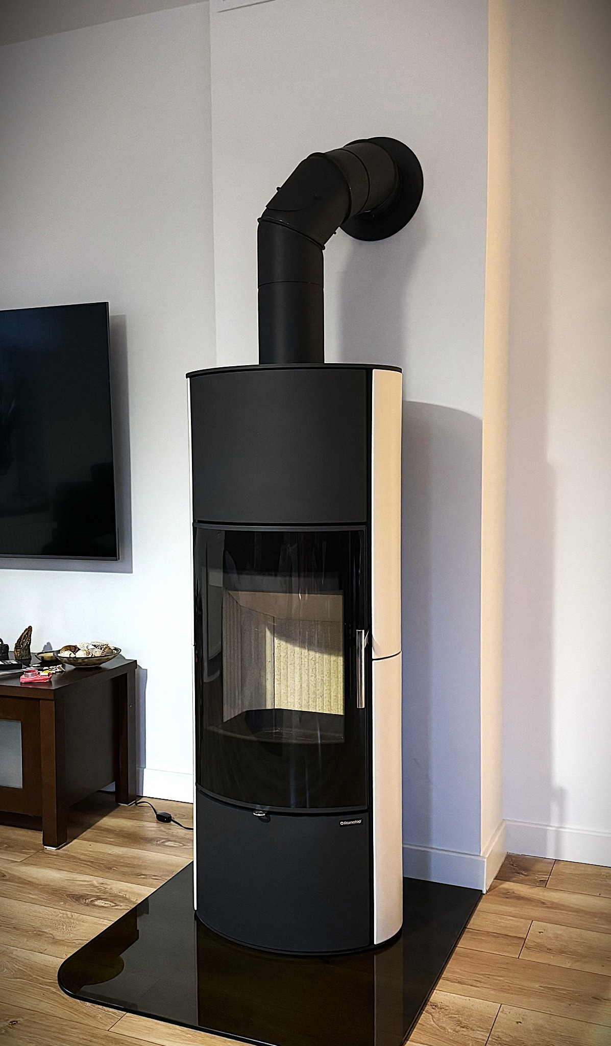 Nowoczesny, czarno-biały kominek wolnostojący z przeszklonym frontem i czarną rurą odprowadzającą spaliny, umieszczony w jasnym pomieszczeniu z parkietem.