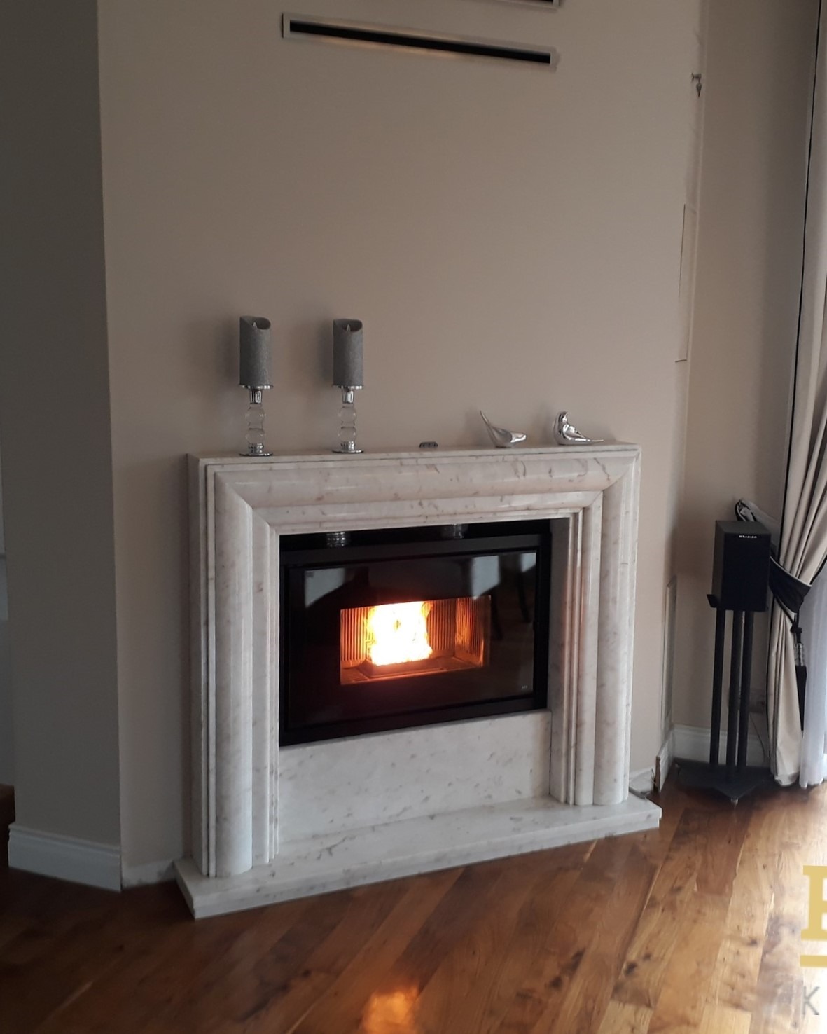 Marmurowy kominek z paleniskiem, płomień wewnątrz, dekoracje na górze, drewniana podłoga, nowoczesny design, jasne wnętrze. Widoczny głośnik na statywie.