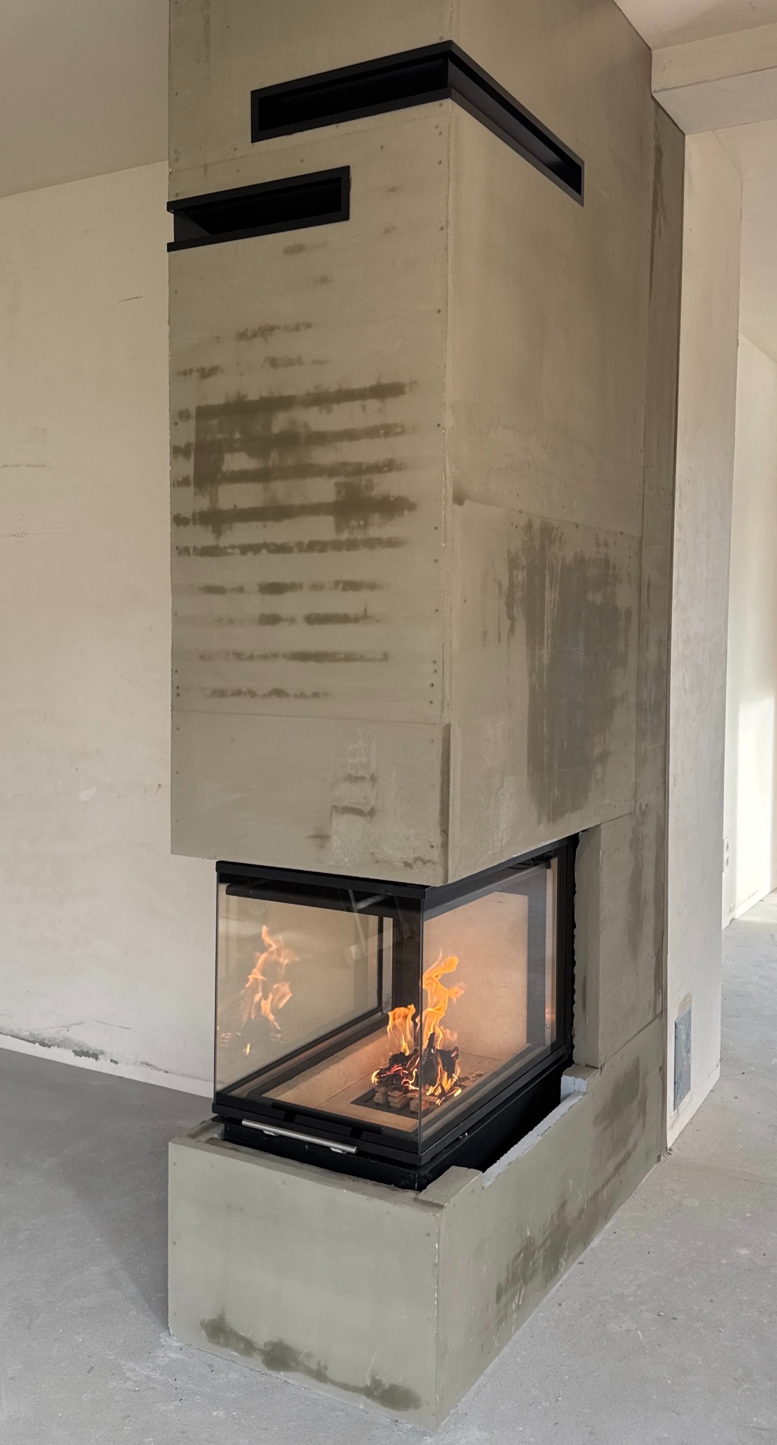 Nowoczesny, surowy kominek z betonu architektonicznego z paleniskiem otoczonym szkłem, płomienie wewnątrz. Minimalistyczny design z czarnymi akcentami.