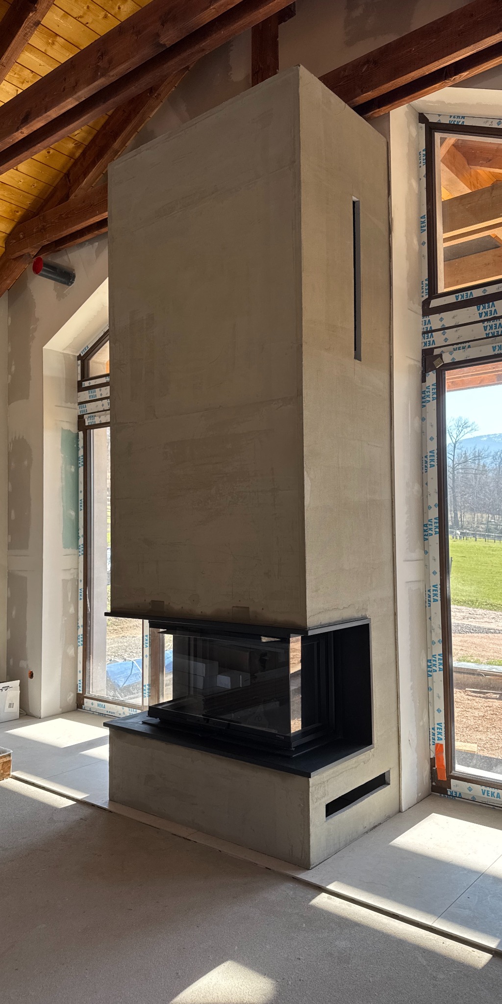Nowoczesny kominek wolnostojący z przeszklonym paleniskiem, surowy beton, w jasnym wnętrzu w trakcie wykończenia. Widoczne drewniane elementy konstrukcyjne dachu.
