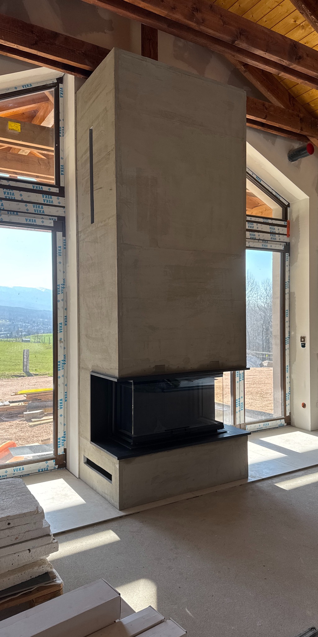 Nowoczesny, surowy kominek z czarnym wkładem, otoczony oknami z widokiem na zielone wzgórza. Wnętrze w trakcie wykończenia, widoczne materiały budowlane.