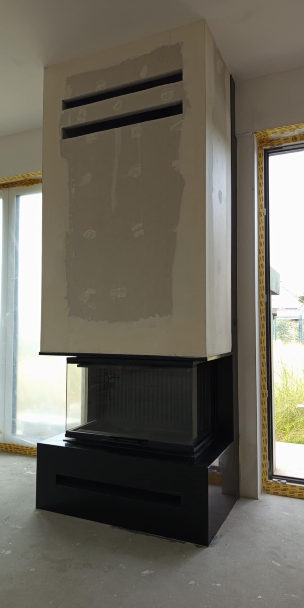 Nowoczesny kominek narożny z szybą i obudową w kolorze czarnym, w trakcie montażu w Oławie. Widoczne otwory wentylacyjne w obudowie i surowy stan wykończenia ściany.