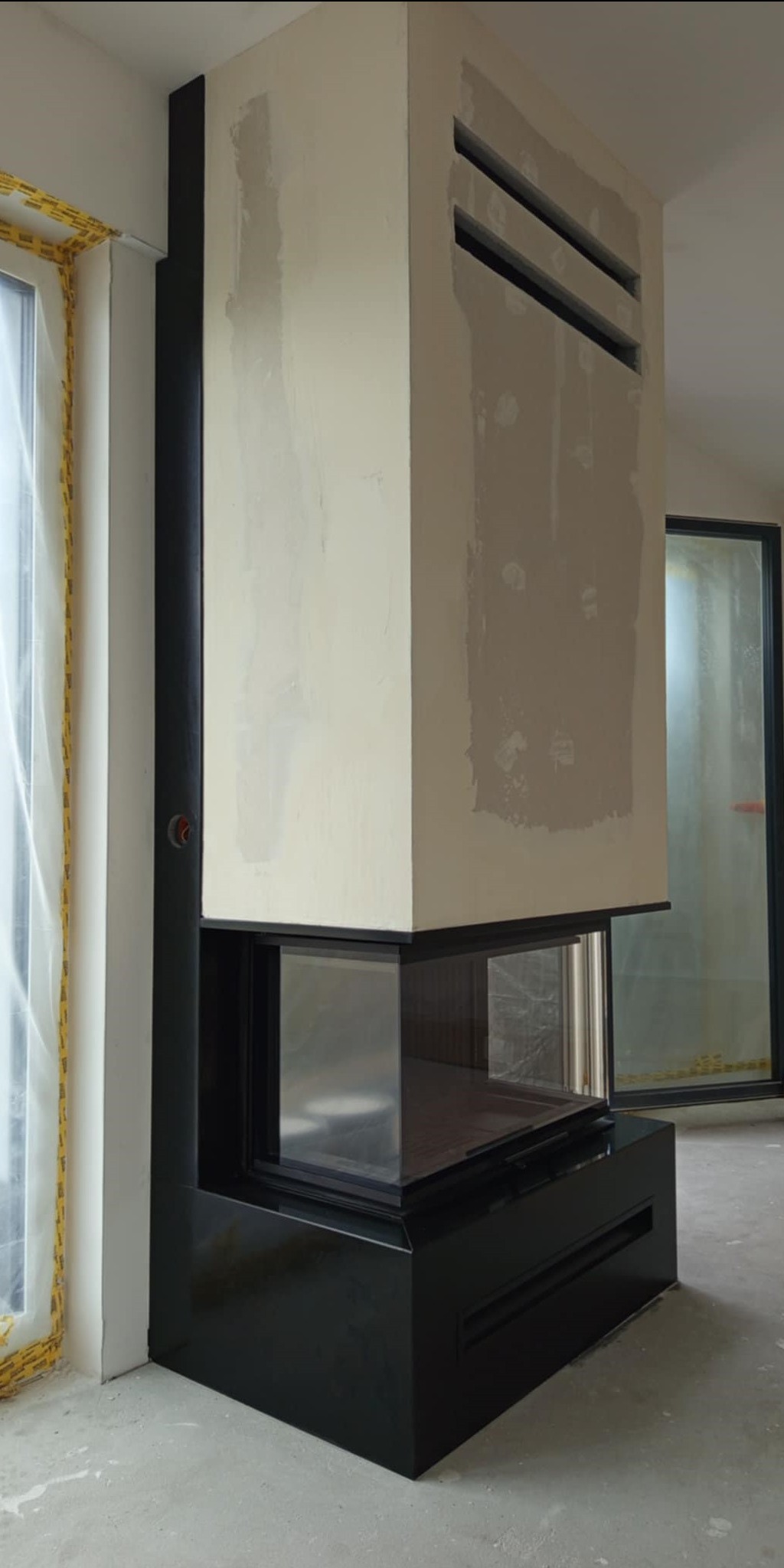 Nowoczesny, narożny kominek z czarną obudową i przeszklonym paleniskiem, w surowym wnętrzu. Widoczne niedokończone wykończenia ścian i okna zabezpieczone folią.