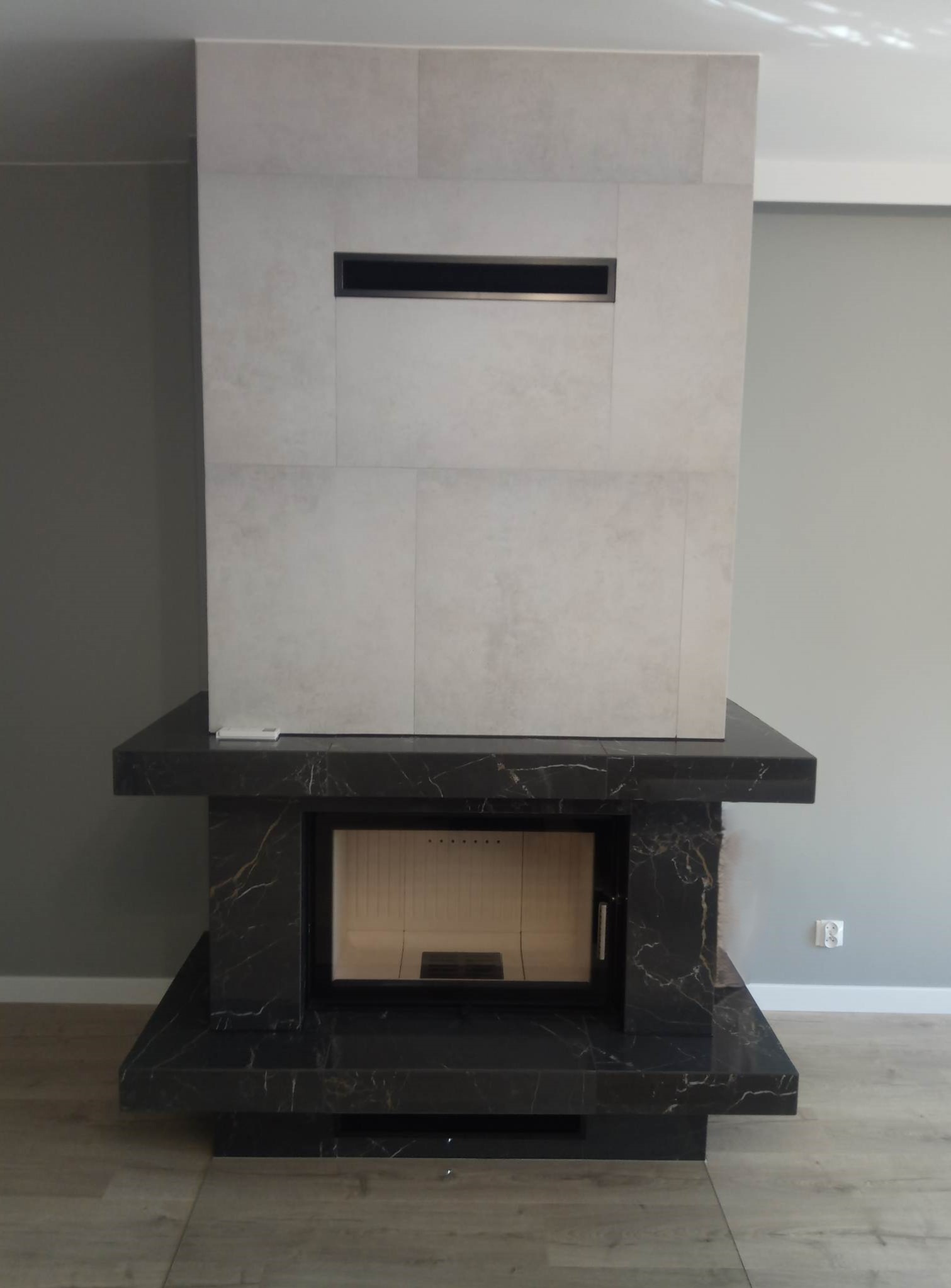 Nowoczesny kominek obudowany płytami w odcieniach szarości i czerni z marmurkowym wzorem, z poziomym wlotem powietrza i zamkniętym paleniskiem.